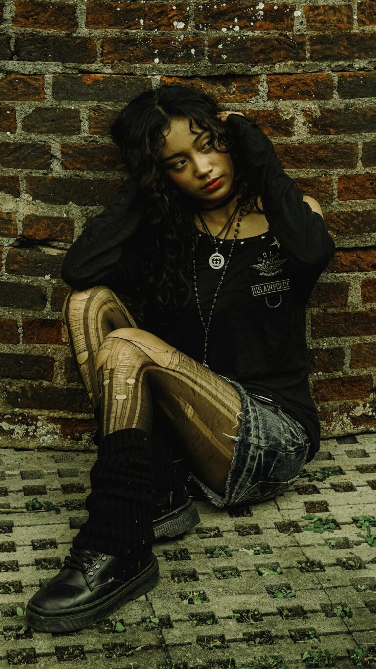 Moody portrait of a young woman in stylish clothing seated against a textured brick wall.