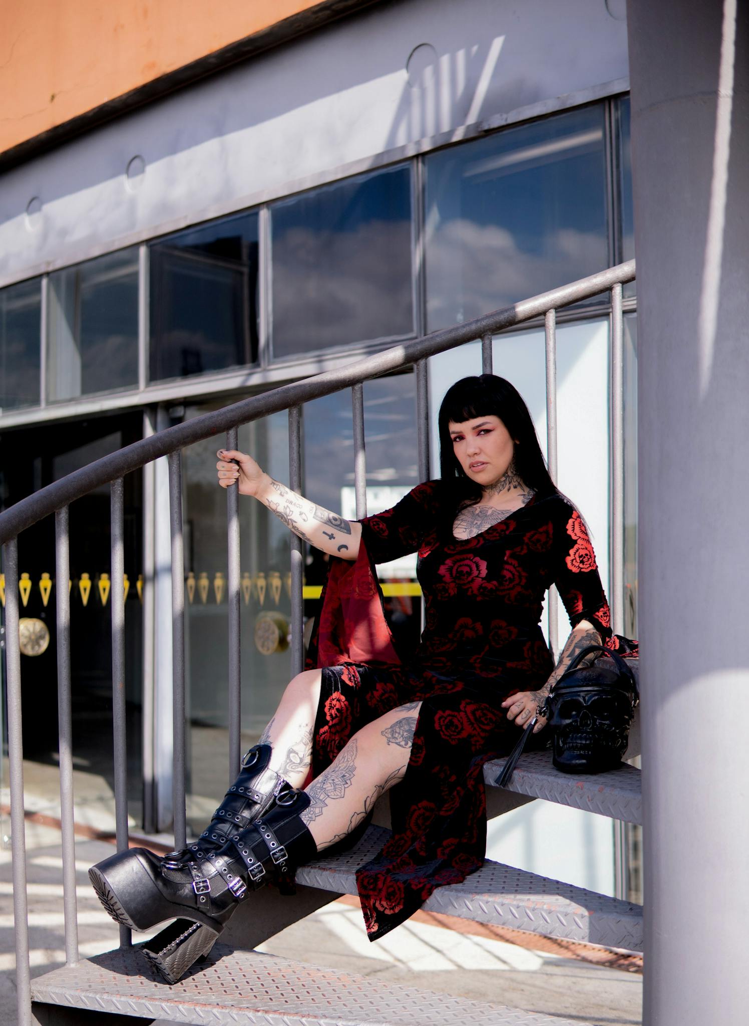 Tattooed woman in gothic attire sitting on spiral stairs outdoors.